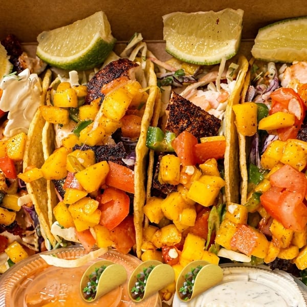 Four fish tacos with mango salsa and lime wedges in a cardboard box with dipping sauces.
