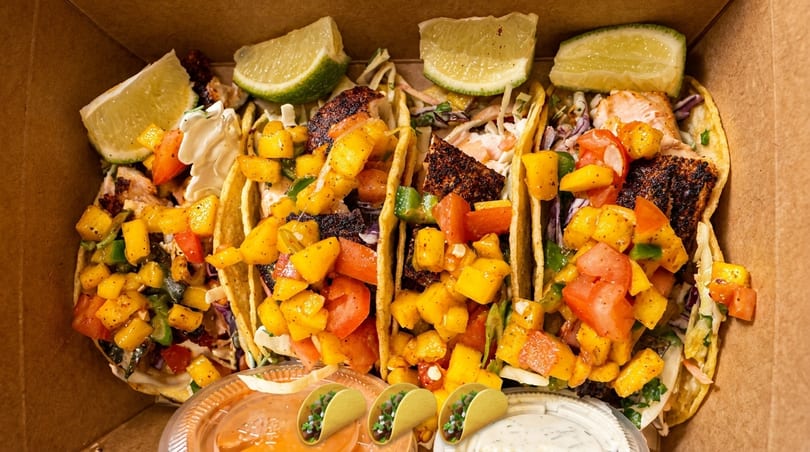 Four fish tacos with mango salsa and lime wedges in a cardboard box with dipping sauces.