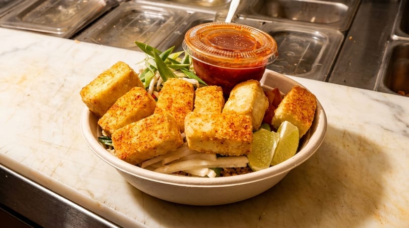 A bowl of crispy fried tofu, green onions, lime wedges, and a side of chili sauce.