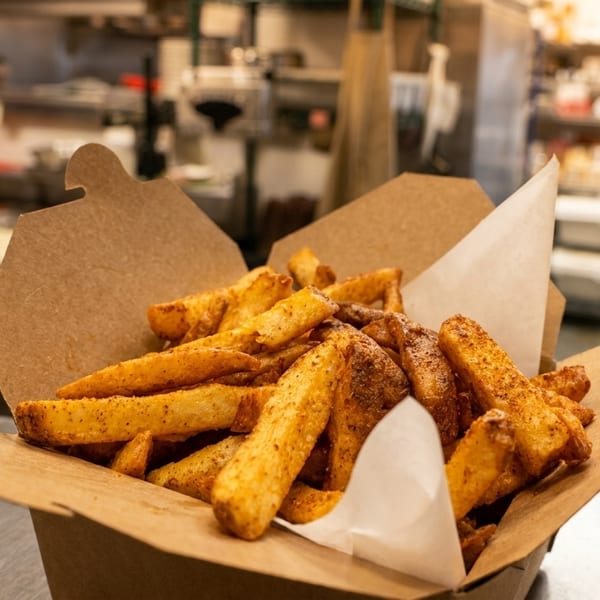 Seasoned french fries in a brown paper container on a metal counter in a commercial kitchen.