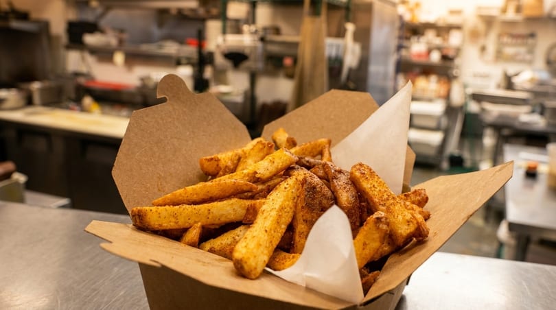 Seasoned french fries in a brown paper container on a metal counter in a commercial kitchen.